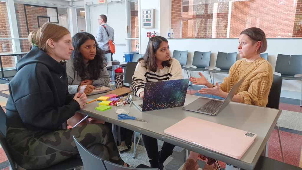 Three students seated at a table listen to a person explaining a project.