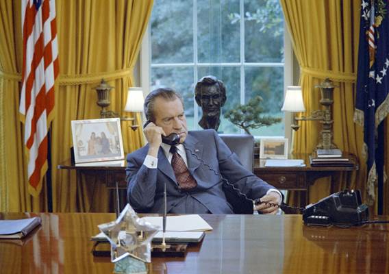 President Richard Nixon, seated at his desk in the Oval Office, speaks on the phone.