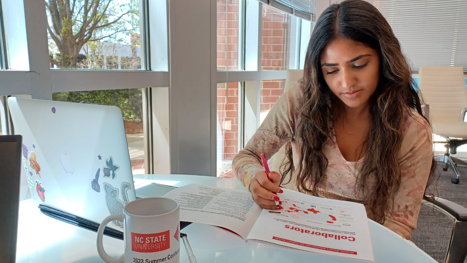 A student with long dark hair holds a pen and reads the impact report. The student's computer and a mug are also on the table. Windows next to the table show brick buildings and trees.