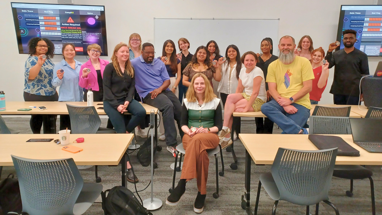 A professor is seated with a dozen students gathered behind her, smiling.