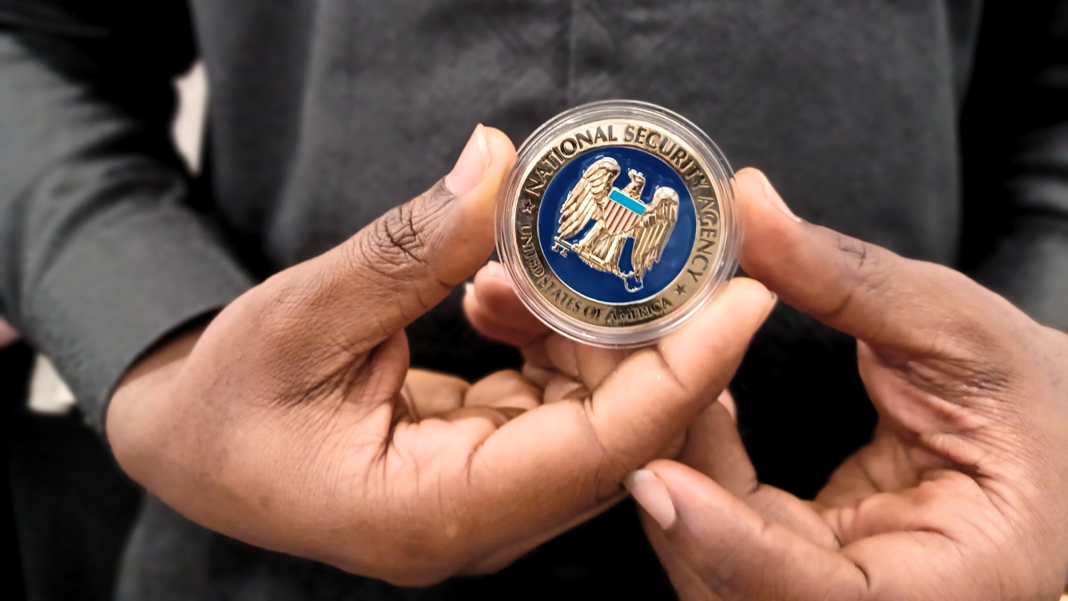 Close up of two hands holding an enamel challenge coin decorated with the National Security Agency logo.