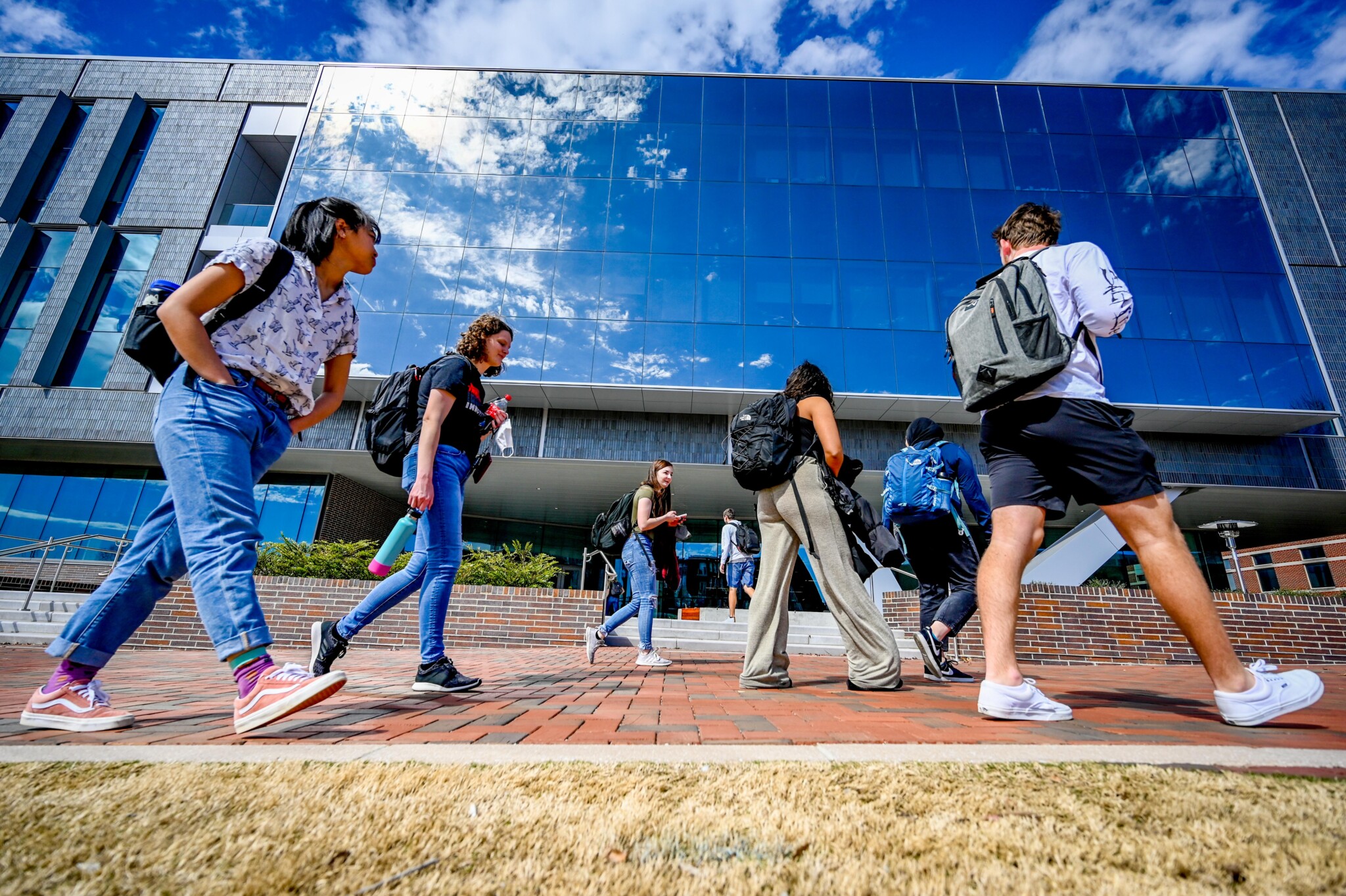 Wide angle shot of college students on a brick walkway, with blue sky reflecting off the windows in the building behind them.